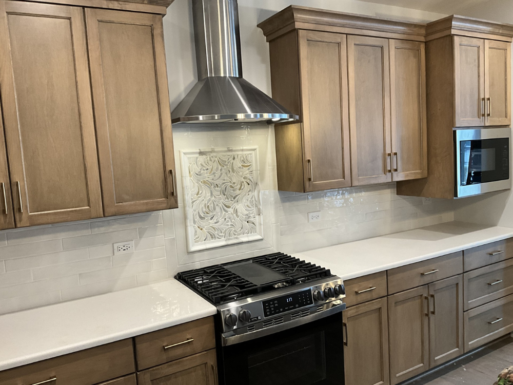 A modern kitchen with light brown cabinets, a stainless steel range hood and stove, and white countertops and backsplash. A microwave is installed above the cabinets on the right.